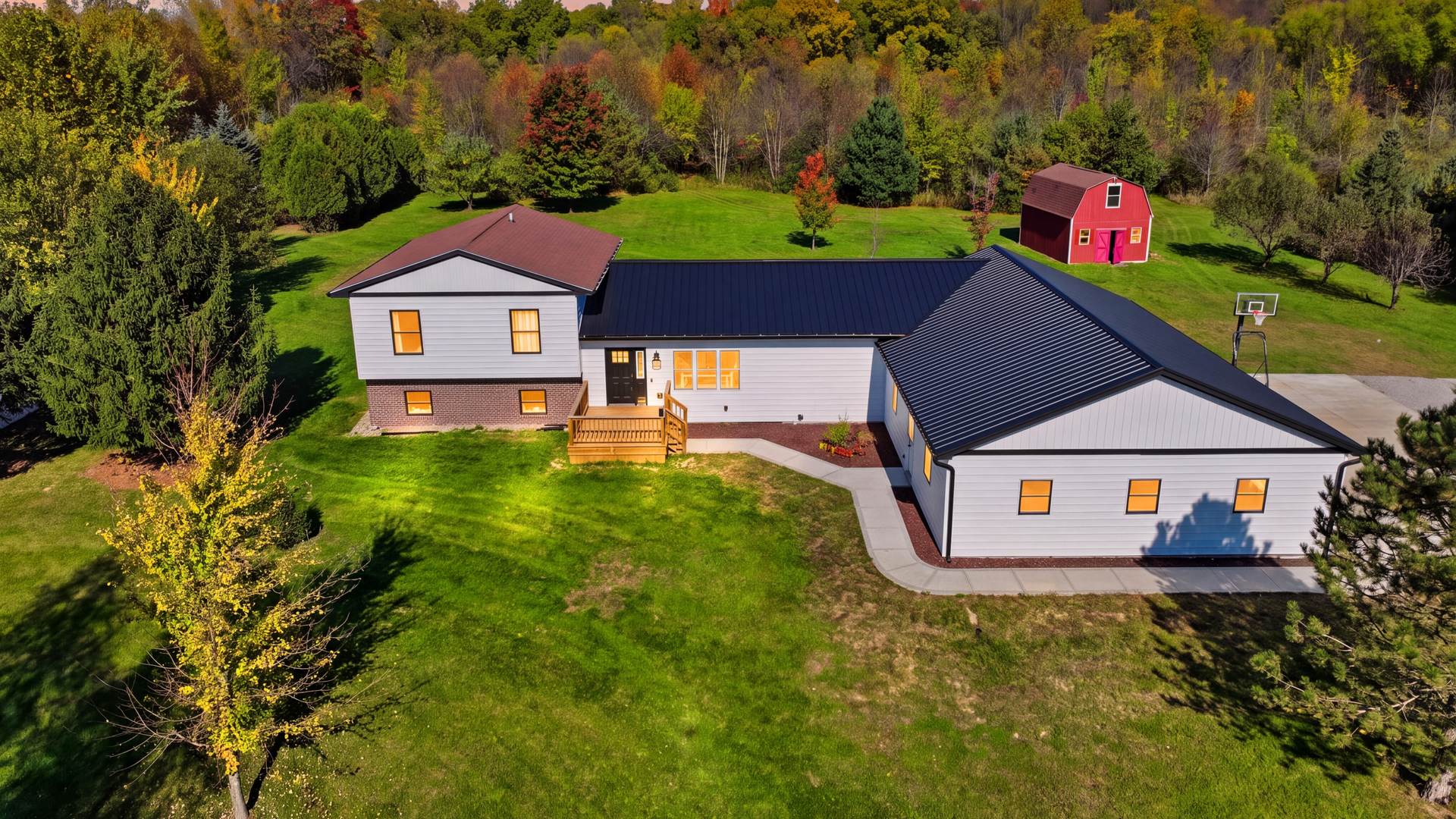 Twilight aerial of a white farmhouse with glowing windows and a red barn on a wooded lot